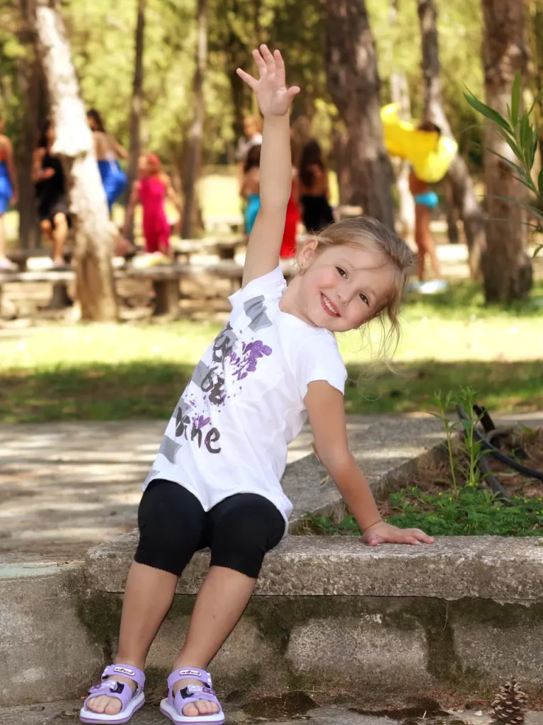 Enfant souriante assise, levant la main dans un parc.